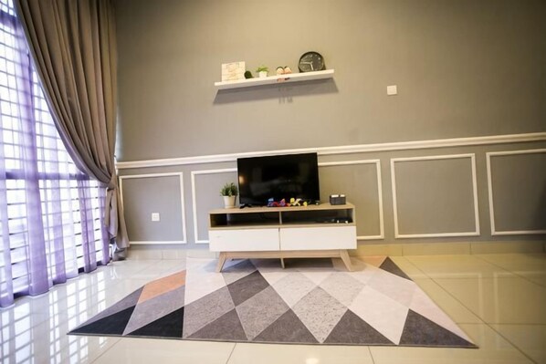 Una sala de estar con una televisión de pantalla plana sobre un mueble blanco, un tapete estampado y una ventana con cortinas.