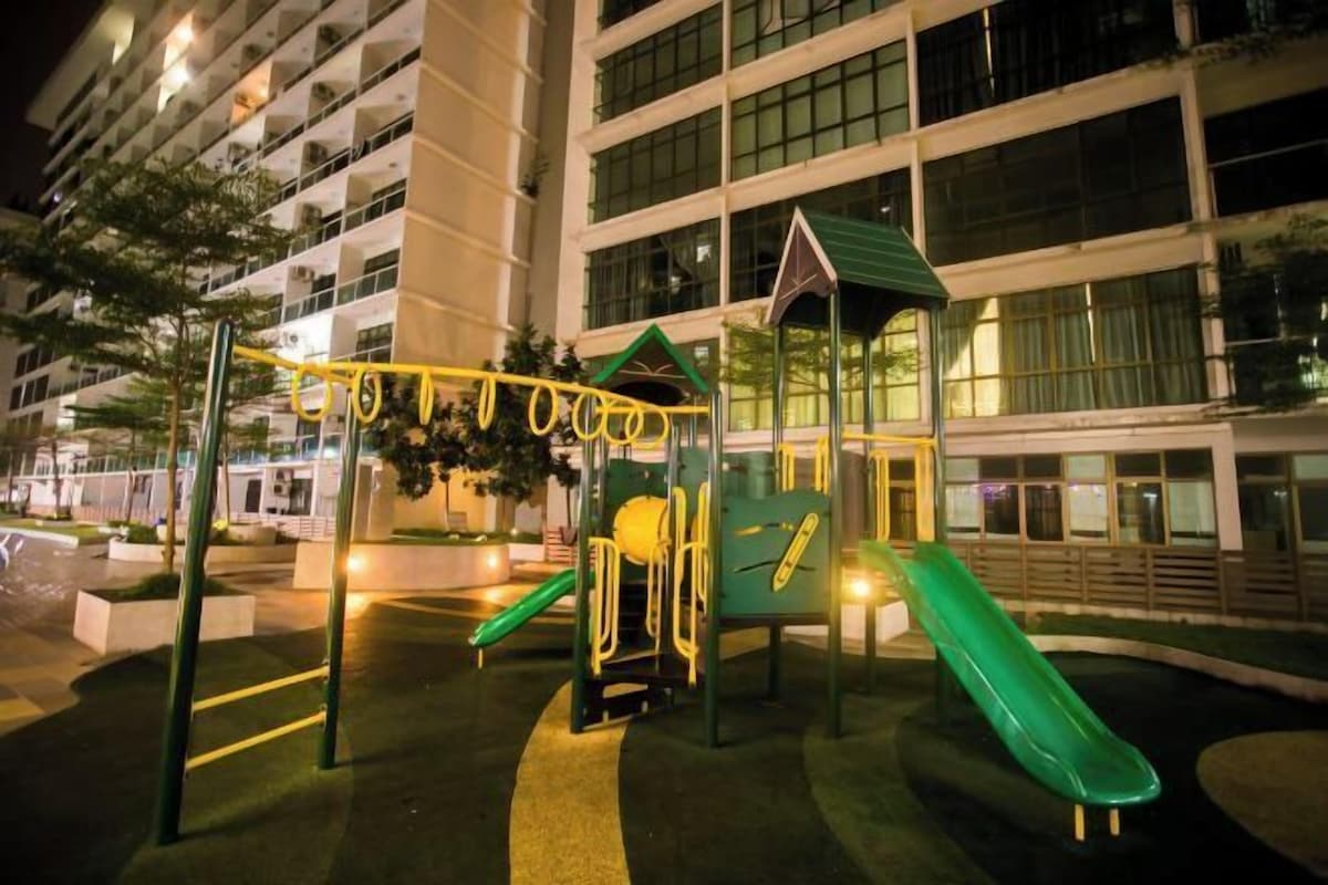 A playground with a slide and climbing structures, surrounded by a building with large windows and a green roof.