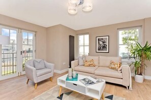 A spacious living room with a sofa, armchair, and coffee table, large windows, and a potted plant. - Modern central 2 bedroom apartment with parking (Edinburgh)
