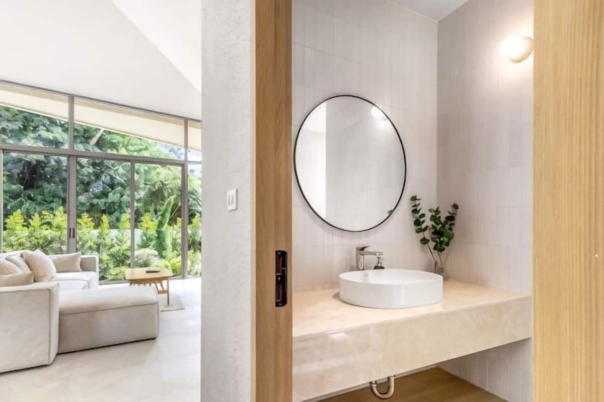 Un baño moderno con un espejo redondo, un lavamanos blanco y vistas a una sala de estar con sofá y ventanas amplias.