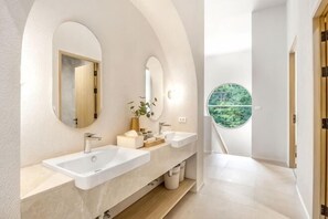 Ein modernes Badezimmer mit zwei Waschbecken, einem Spiegel und Blick auf Grün durch ein rundes Fenster.