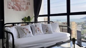 A bed with white bedding and floral pillows, a city view through large windows, and a wall with framed artwork. - Georgetown Urban Suite Family Skypool I SeaView (Jelutong)