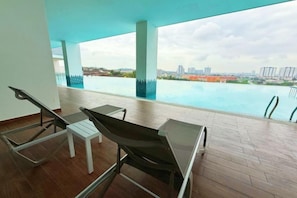A poolside area with lounge chairs and a small table, overlooking a body of water with a cityscape in the background. - Nexus Kajang by Luvit Stay (Kajang)