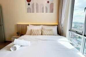 Un letto rifatto con lenzuola bianche e cuscini decorativi, un comodino con una lampada e vista sulla città attraverso un'ampia finestra.