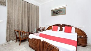 Habitación de hotel con una cama grande, cabecero de madera, un escritorio con una silla, una mesita de noche y un ventanal con cortinas.