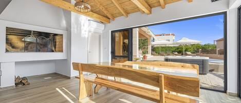 Una cocina moderna con un banco de madera, un horno de ladrillo y un gran ventanal corredero que da a un área exterior con piscina y sombrillas.