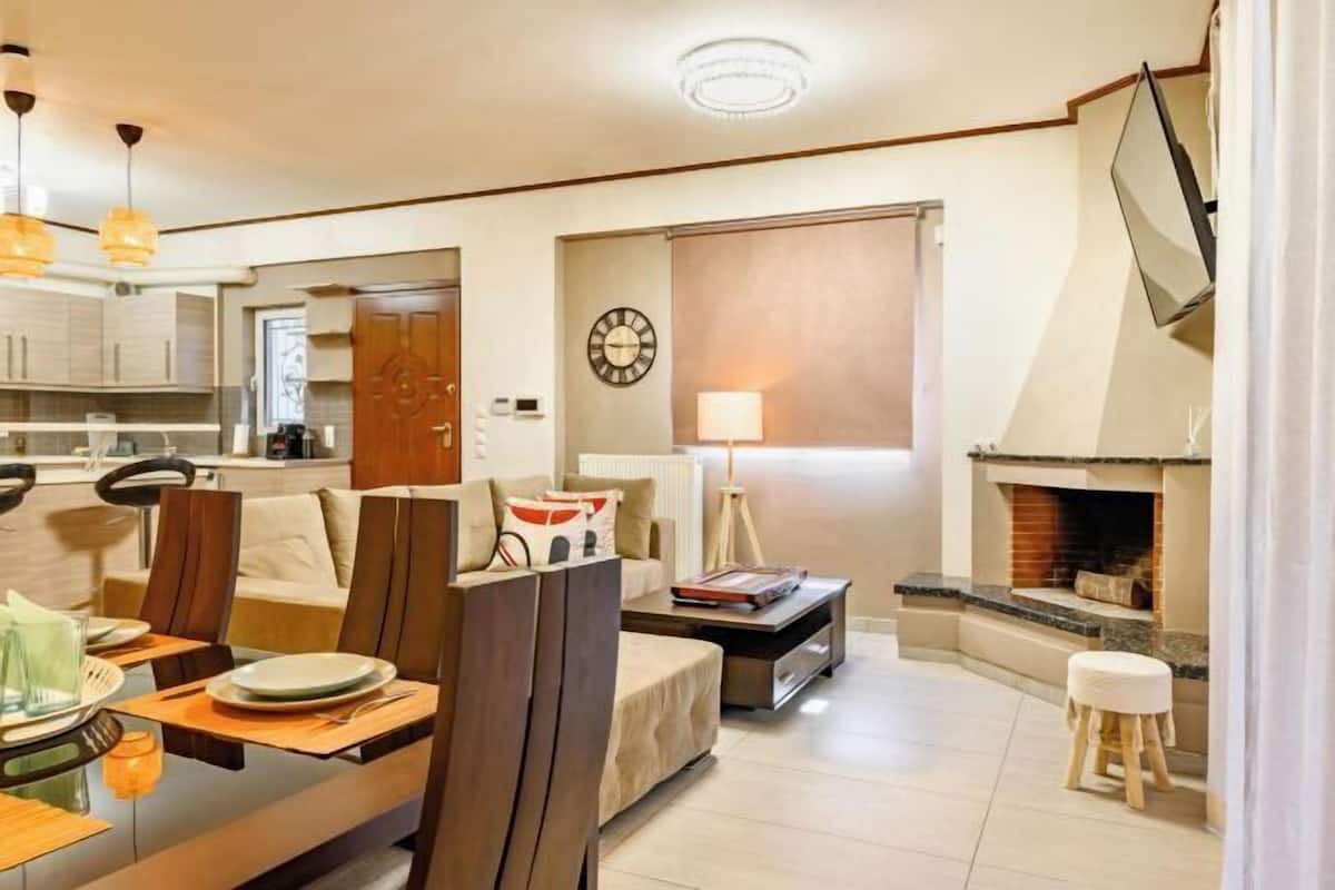 Un salon moderne avec une cheminée, un coin repas avec une table dressée pour deux personnes, et une cuisine en arrière-plan.