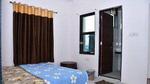 Un dormitorio con cama, cortinas, una ventana, una puerta, un taburete pequeño y un cubo de basura.