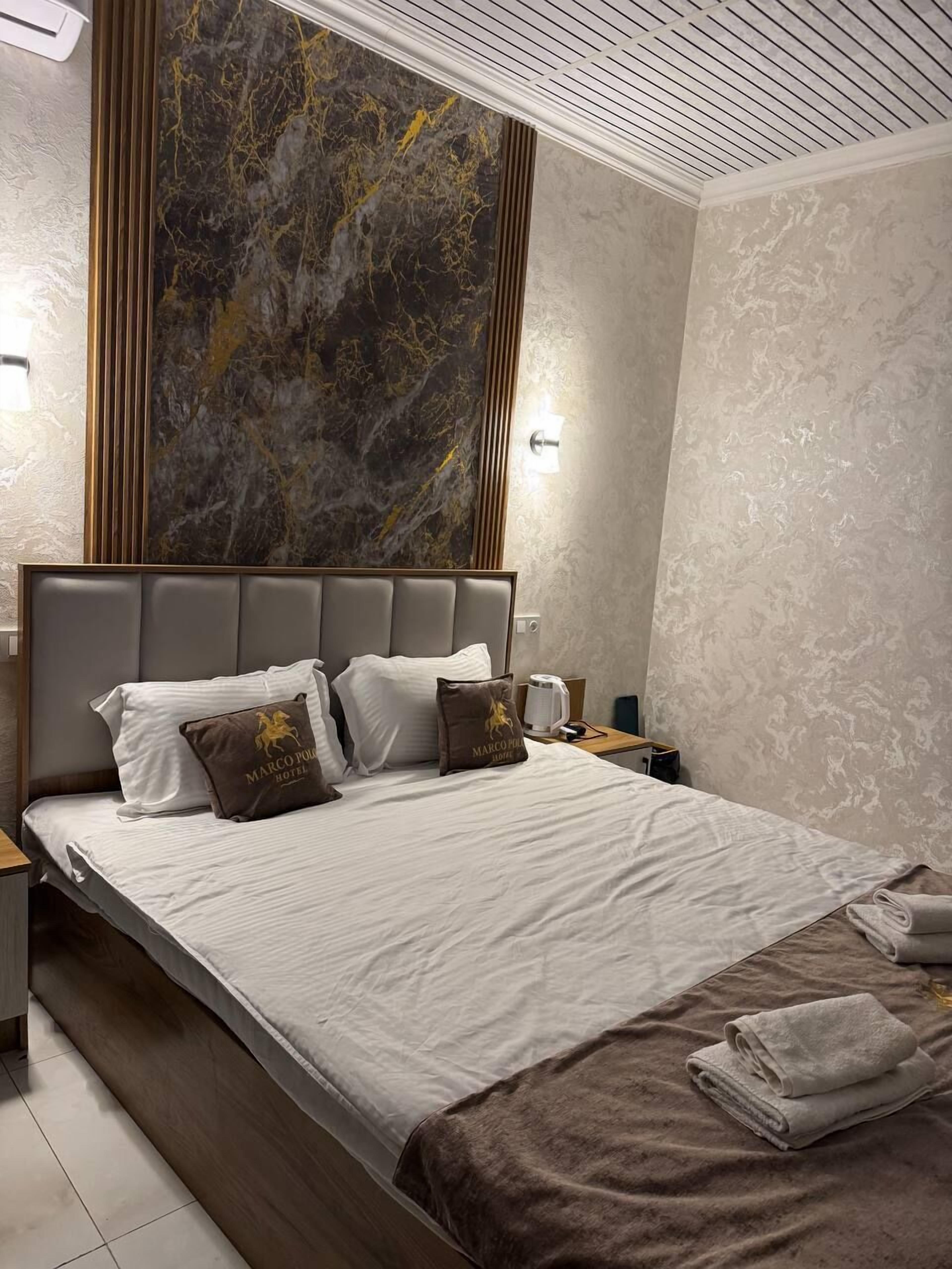 Quarto de hotel com uma cama grande, parede com detalhes em mármore e design moderno no teto.