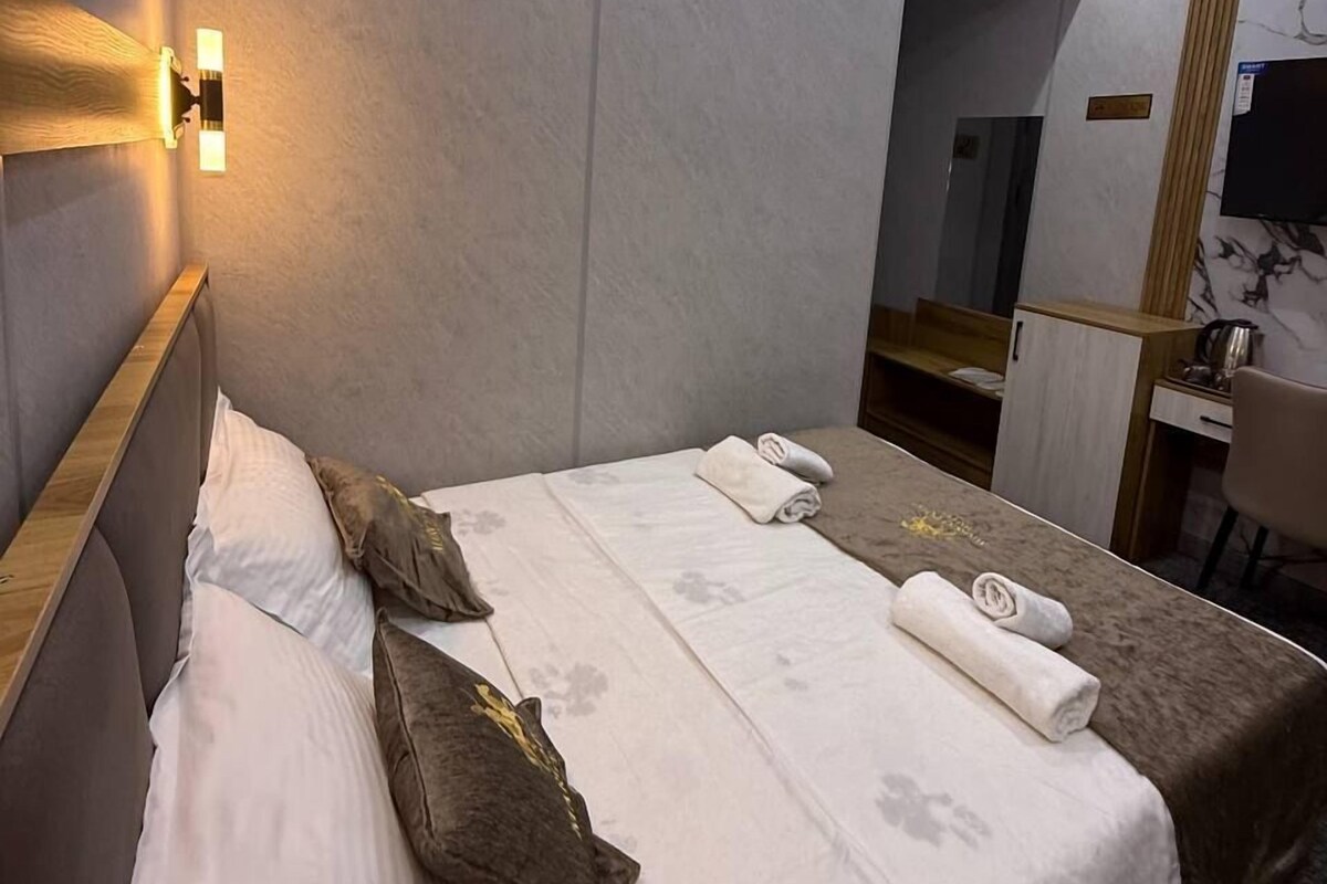 A modern hotel room with a large bed, a bedside table, a mirror, and a desk with a chair.