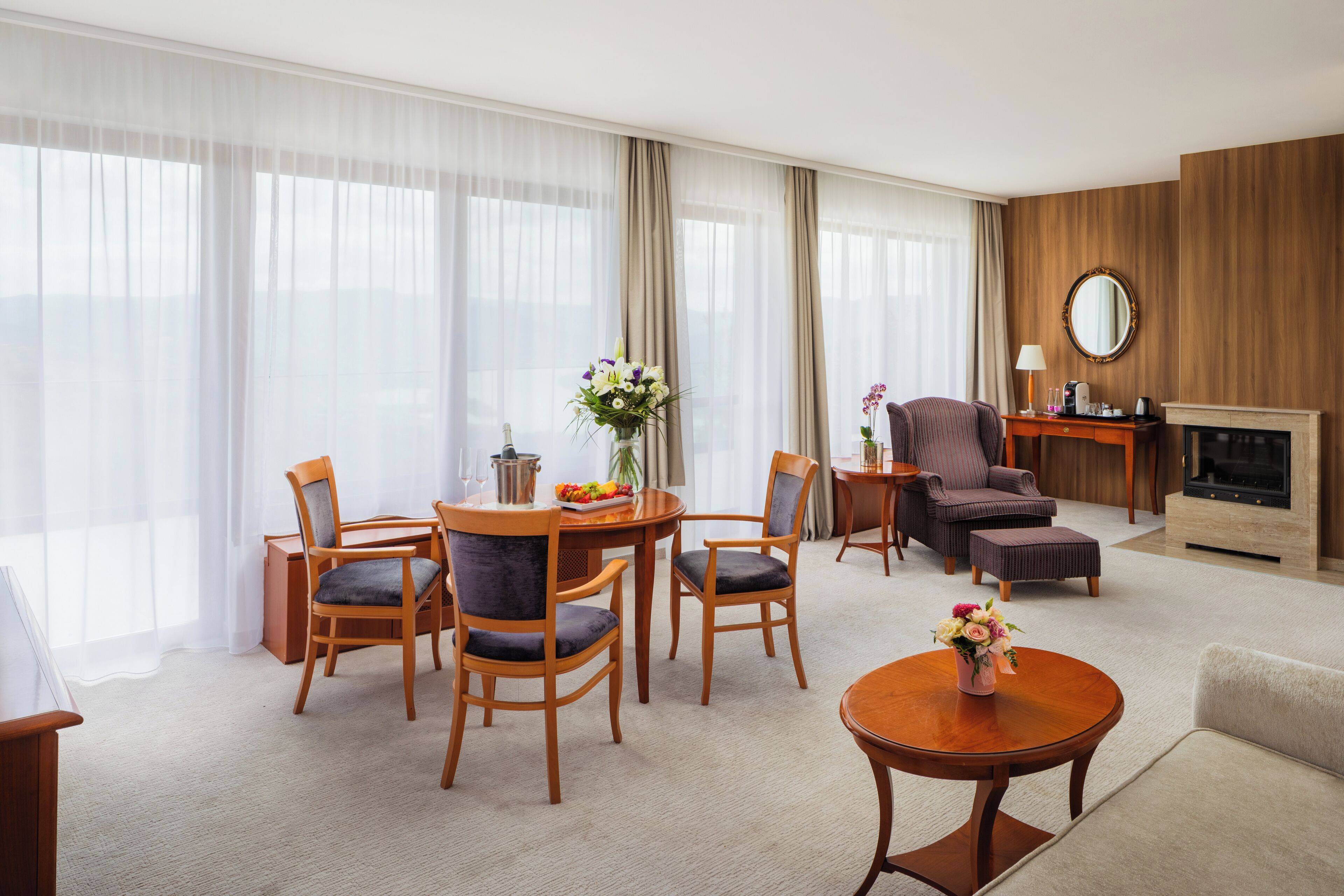 A hotel room with a dining area, a sofa, a fireplace, and a large window with sheer curtains.