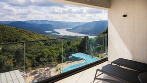 A balcony with a glass railing overlooking a swimming pool and a mountainous landscape. - Silvanus Hotel (Visegrád)