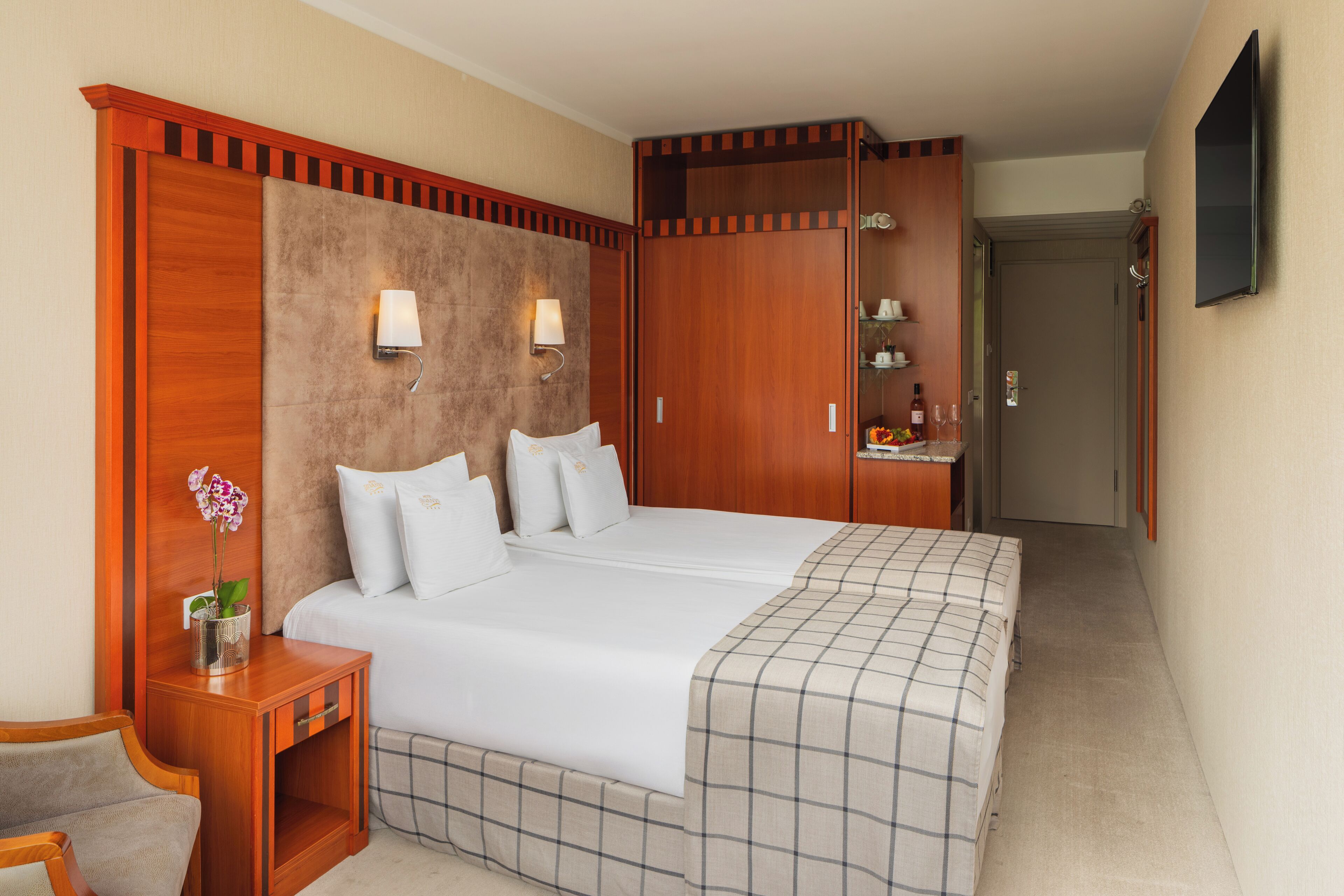Una habitación de hotel con una cama grande, una mesita de noche con una lámpara, una pequeña mesa con un jarrón de flores y un televisor montado en la pared.