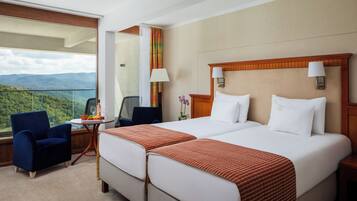 Quarto de hotel com duas camas, vista para montanhas, uma cadeira, uma mesinha com frutas e um abajur.
