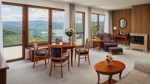A spacious room with a dining area, a sofa, a fireplace, and large windows offering a scenic view. - Silvanus Hotel (Visegrád)