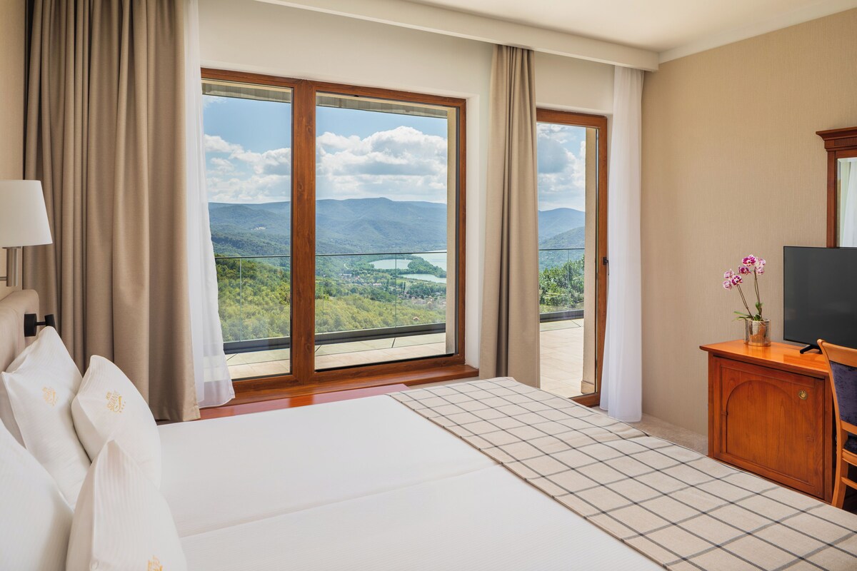 A hotel room with a large bed, a TV, a wooden cabinet, and a view of mountains and a lake.