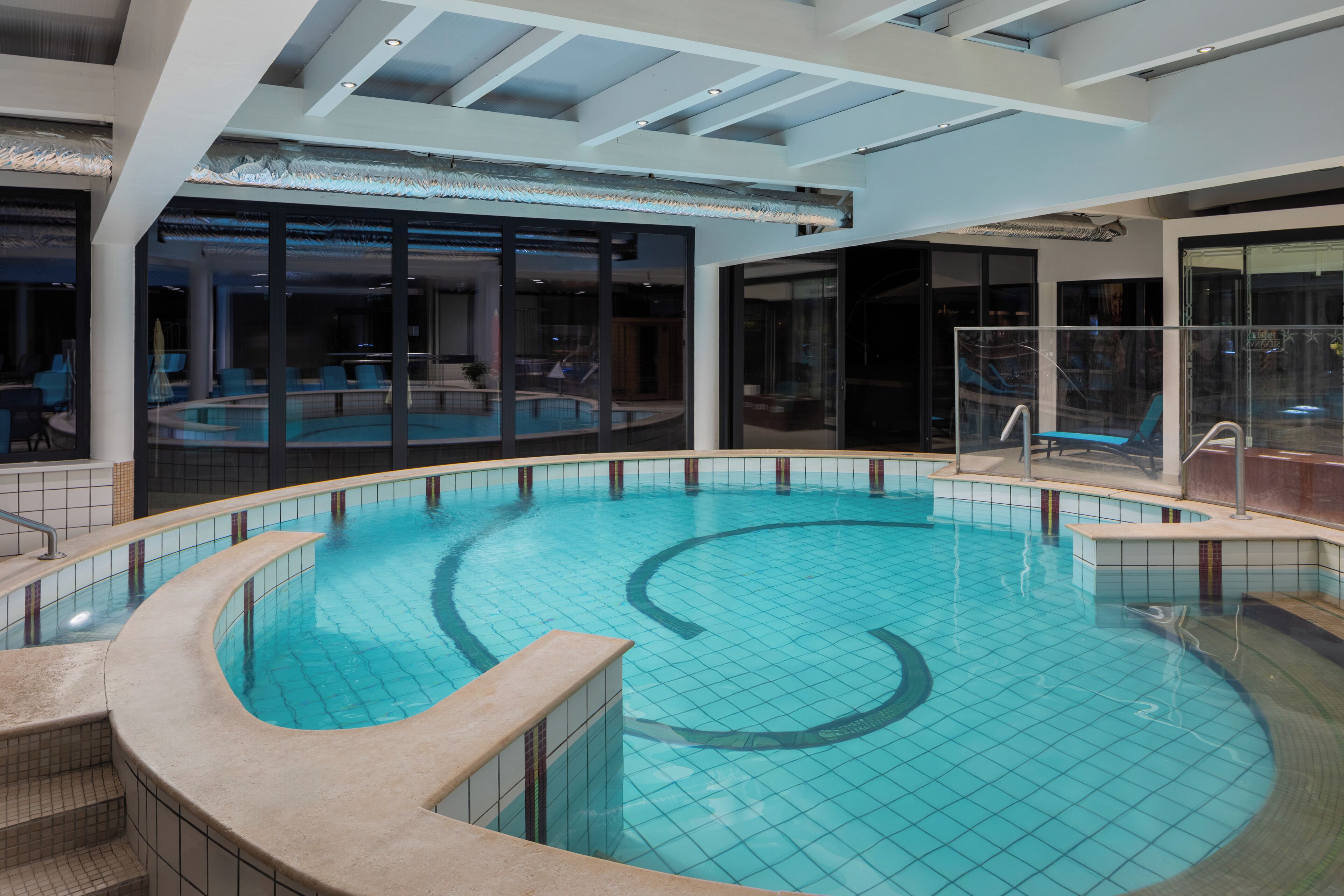 An indoor swimming pool with a curved design, surrounded by glass walls and a modern ceiling structure.