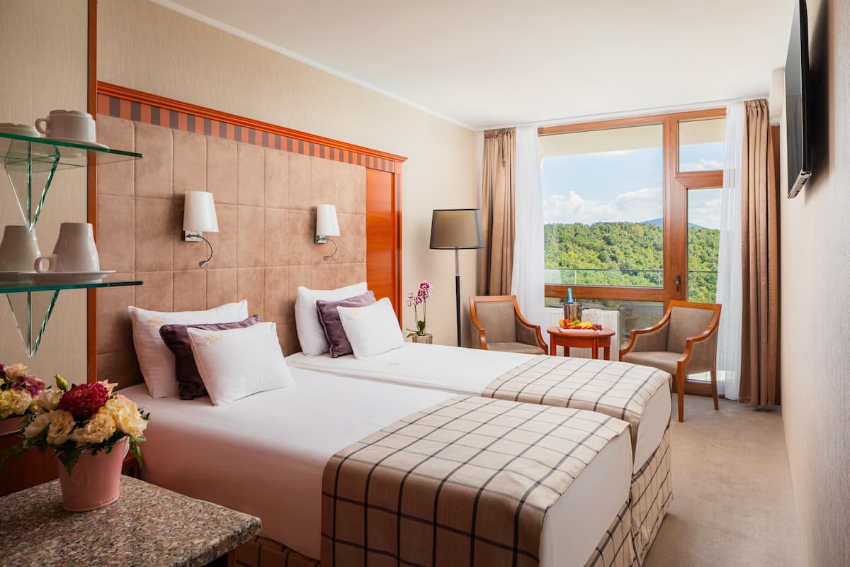 Habitación de hotel con dos camas, zona de estar con mesa y sillas, televisor montado en la pared y ventana con vistas a zonas verdes.