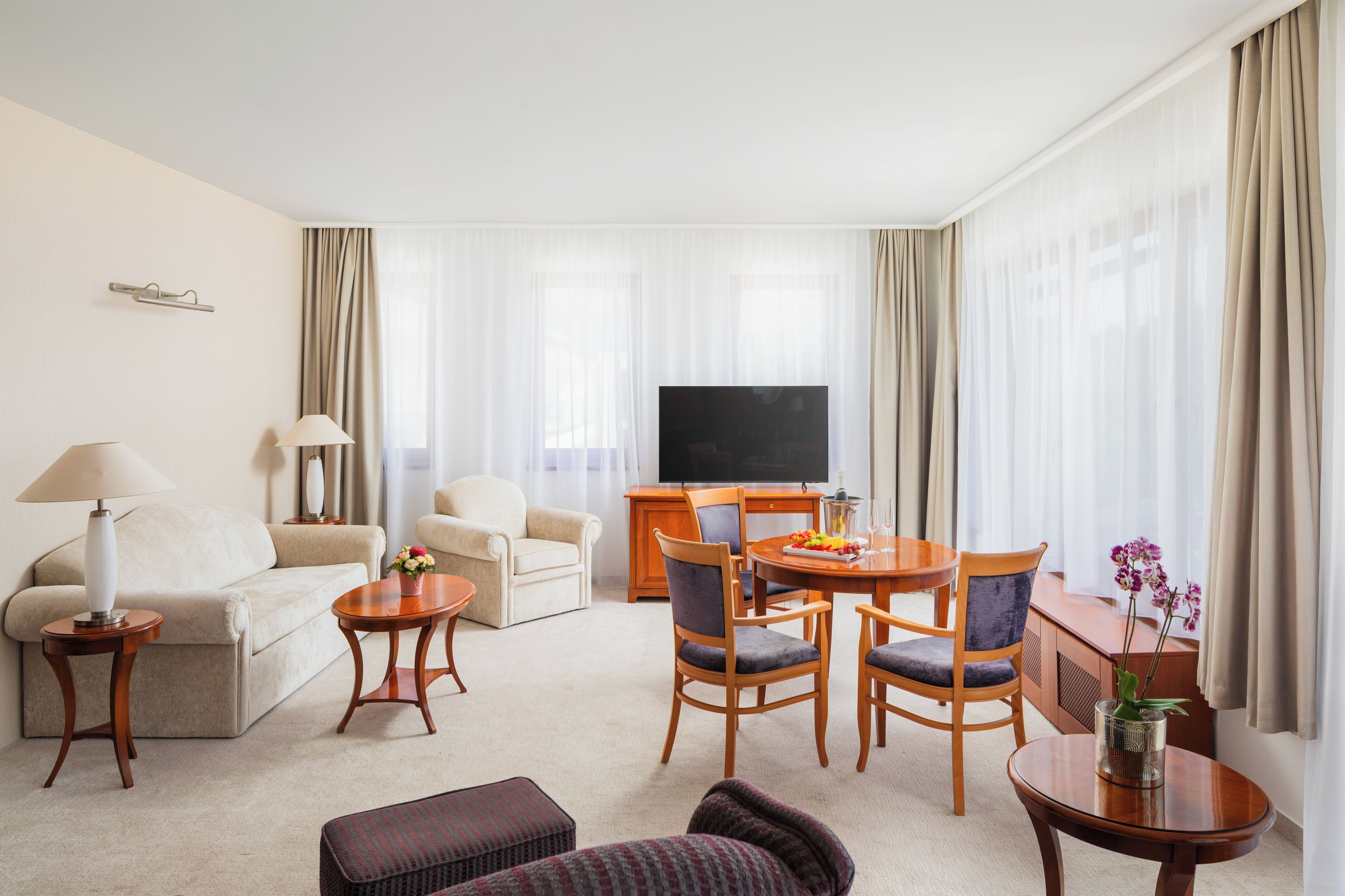 A hotel room with a sofa, a dining table with chairs, a TV, and a small side table with a lamp.