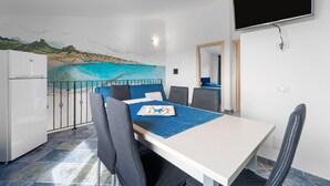 A dining area with a table, chairs, a painting of a beach, a TV mounted on the wall, and a refrigerator. - Villa Le Torri Porto Corallo (Villaputzu)