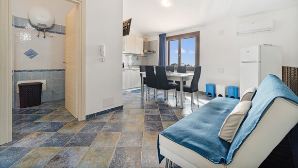A compact kitchen with a dining area, a small trash bin, and a window. - Villa Le Torri Porto Corallo (Villaputzu)
