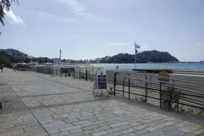 Une promenade en bord de mer, dallée, avec une rambarde en métal, un panneau d’information et un drapeau.