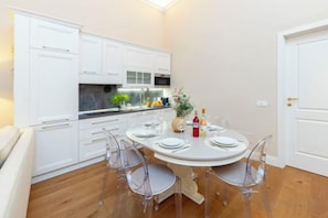 Een kleine keuken met witte kastjes, een eettafel met stoelen en een deur aan de rechterkant.