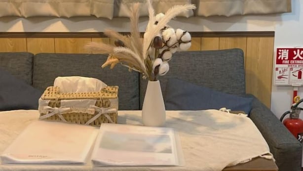 Une pièce comprenant un lit, une table de chevet, un vase avec des fleurs séchées, un panier et un extincteur fixé au mur.
