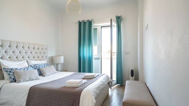 A bedroom with a tufted headboard, a large bed with white and patterned bedding, a bench, teal curtains, and a view through a glass door. - Jambul Apartment, Graça, Lisbon (Lisboa)