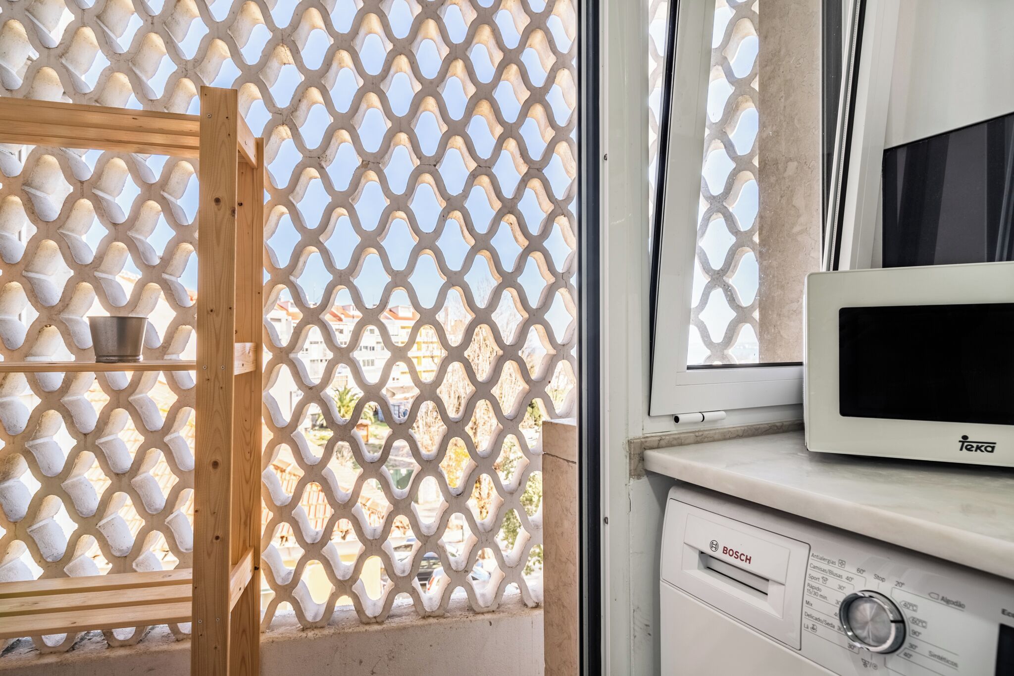 A kitchen with a microwave and a Bosch washing machine, a wooden shelf, and a lattice window.