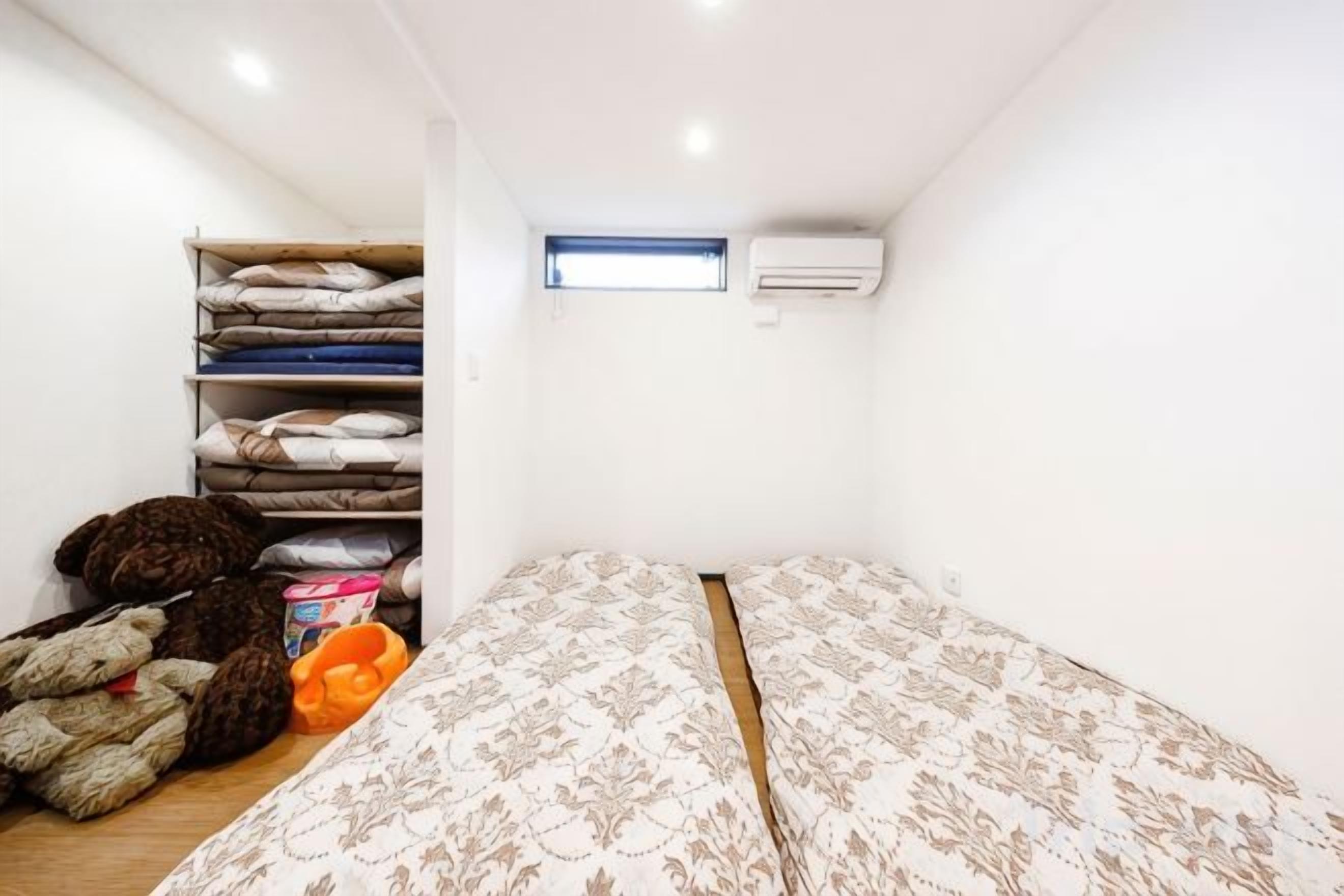 Een eenvoudige kamer met een bed, een plank met opgevouwen dekens, en een airconditioning.