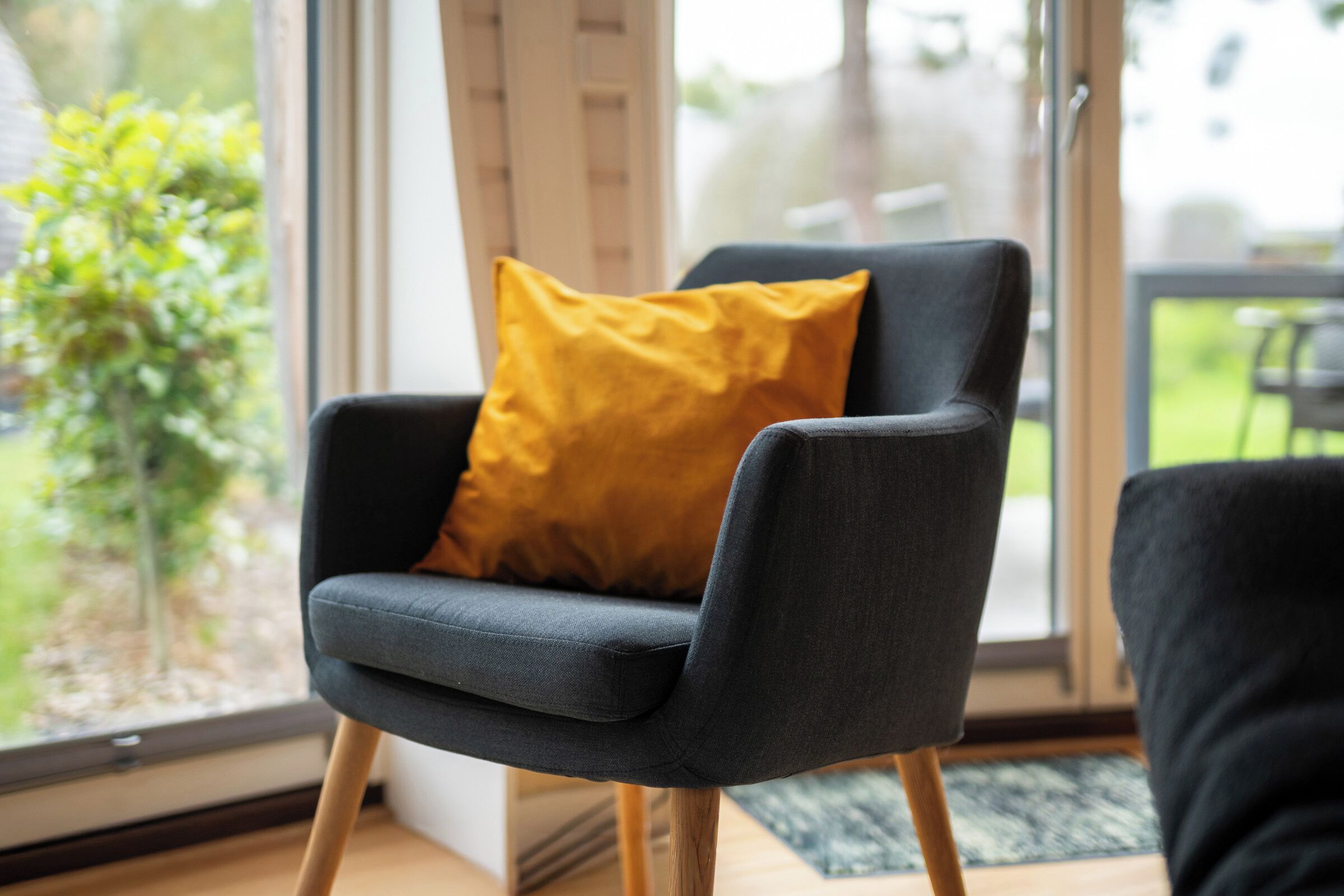 Ein moderner Sessel mit Holzfuß und einer Sitzkissen, platziert in der Nähe eines großen Fensters mit Blick auf Grünfläche.