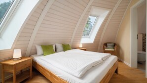 A bedroom with a sloped ceiling, a skylight, a bed with white bedding, a nightstand with a lamp, and a wicker chair. - Vacation home "Kirana" on Lake Tollensesee in Penzlin Wustrow (Penzlin)