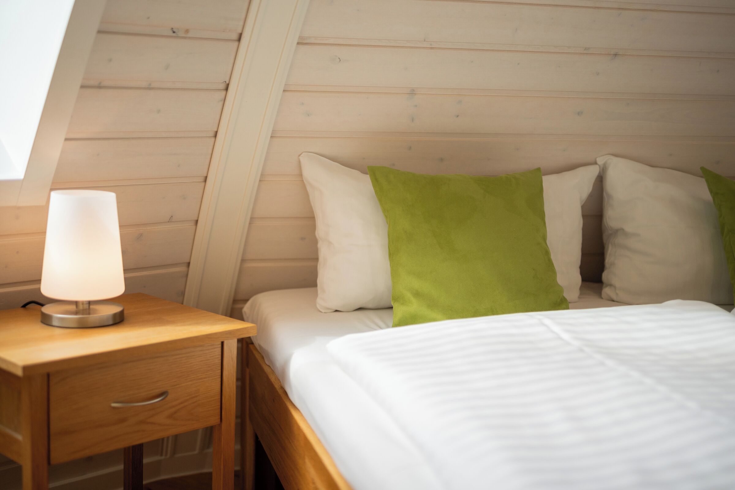 A bedroom with a wooden bed, white bedding, a green pillow, and a bedside lamp on a wooden nightstand.