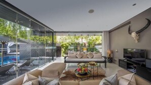 A modern living room with a glass wall, a sofa set, a coffee table with fruit, a TV, and a bull skull decoration. - Villa Balimu Seminyak (Bali)