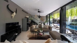 A modern living room with a sofa, TV, and a glass wall leading to a pool area. - Villa Balimu Seminyak (Bali)