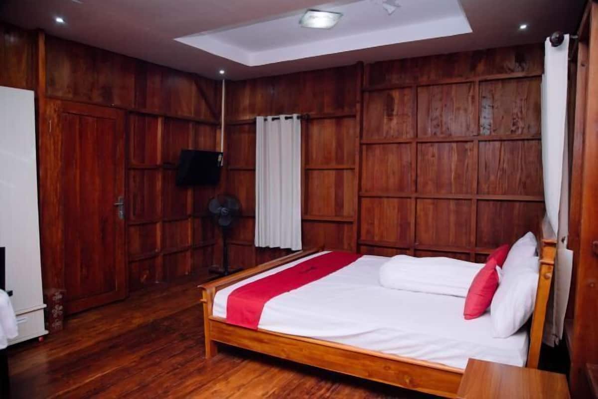 A bedroom with a wooden floor, a bed with white and red bedding, a ceiling fan, and wooden panel walls.