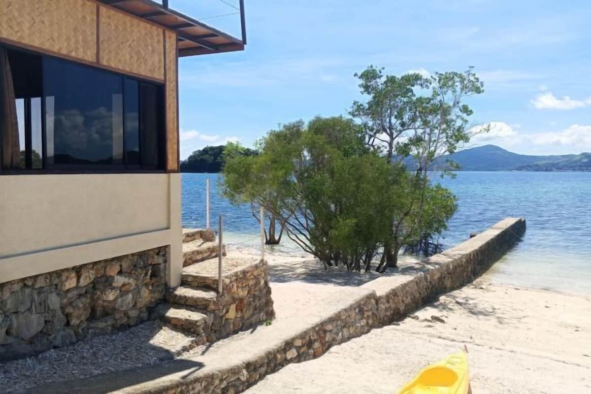 A coastal building with a stone staircase leading to a sandy beach, a kayak, and a tree by the water.
