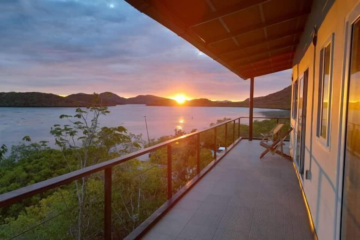 A balcony with a view of a sunset over a body of water, surrounded by greenery.