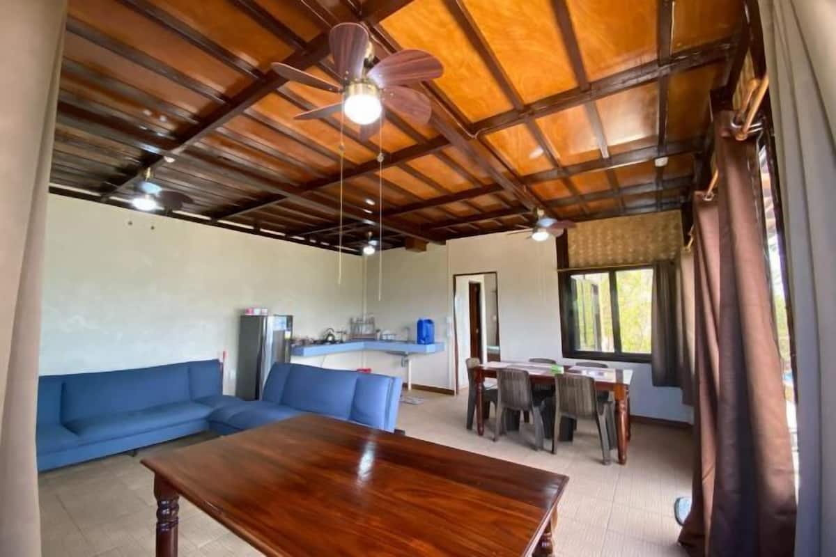 A spacious living area with a wooden ceiling, a blue sofa, a dining table, and a kitchen in the background.