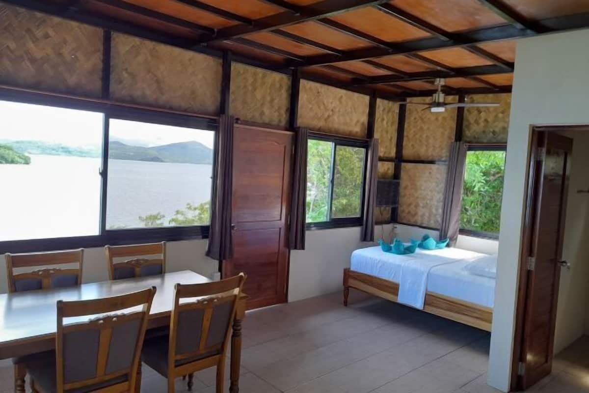 A room with a bed, a table, and chairs. There is a window with a view of a body of water and hills.