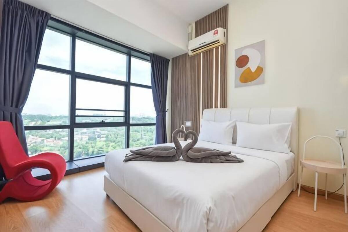A hotel room with a large bed, a red chair, a white chair, and a view of the city through large windows.