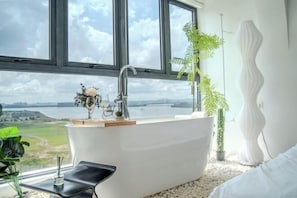 A modern bathroom with a freestanding bathtub, a large window offering a view of a body of water and greenery, and a white vase with flowers on the windowsill.