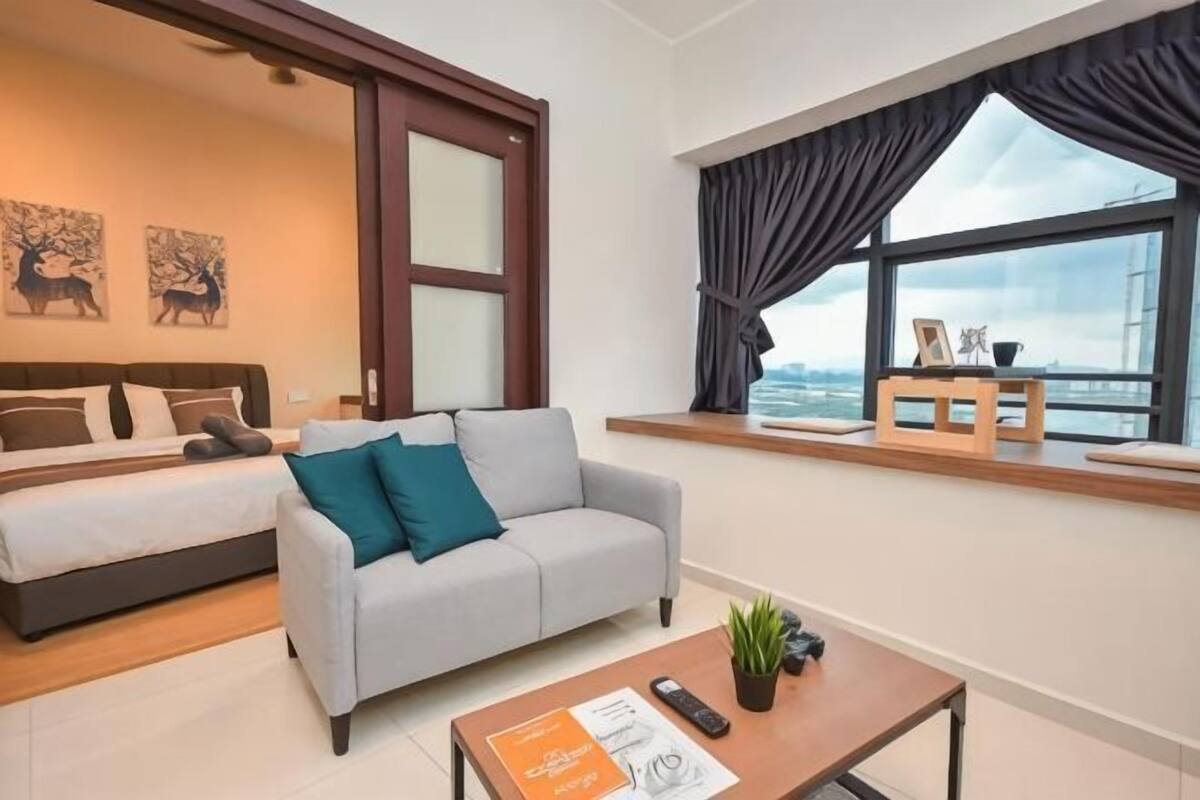 Quarto de hotel moderno com uma cama, um sofá cinza, uma mesa de centro com controle remoto e vista da cidade através de grandes janelas.