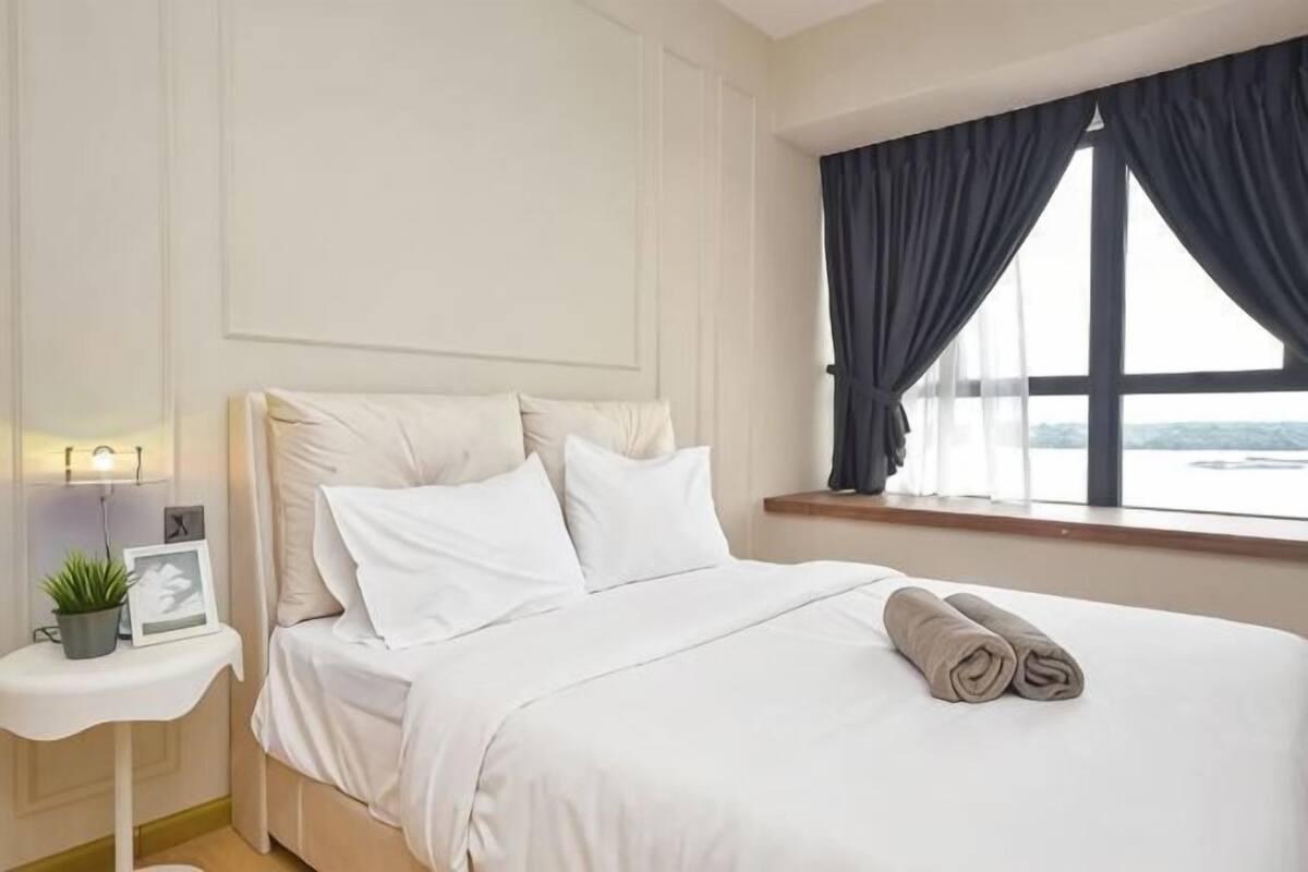 Uma cama bem arrumada com lençóis brancos, uma mesa de cabeceira com abajur e uma pequena planta, e uma janela com cortinas.