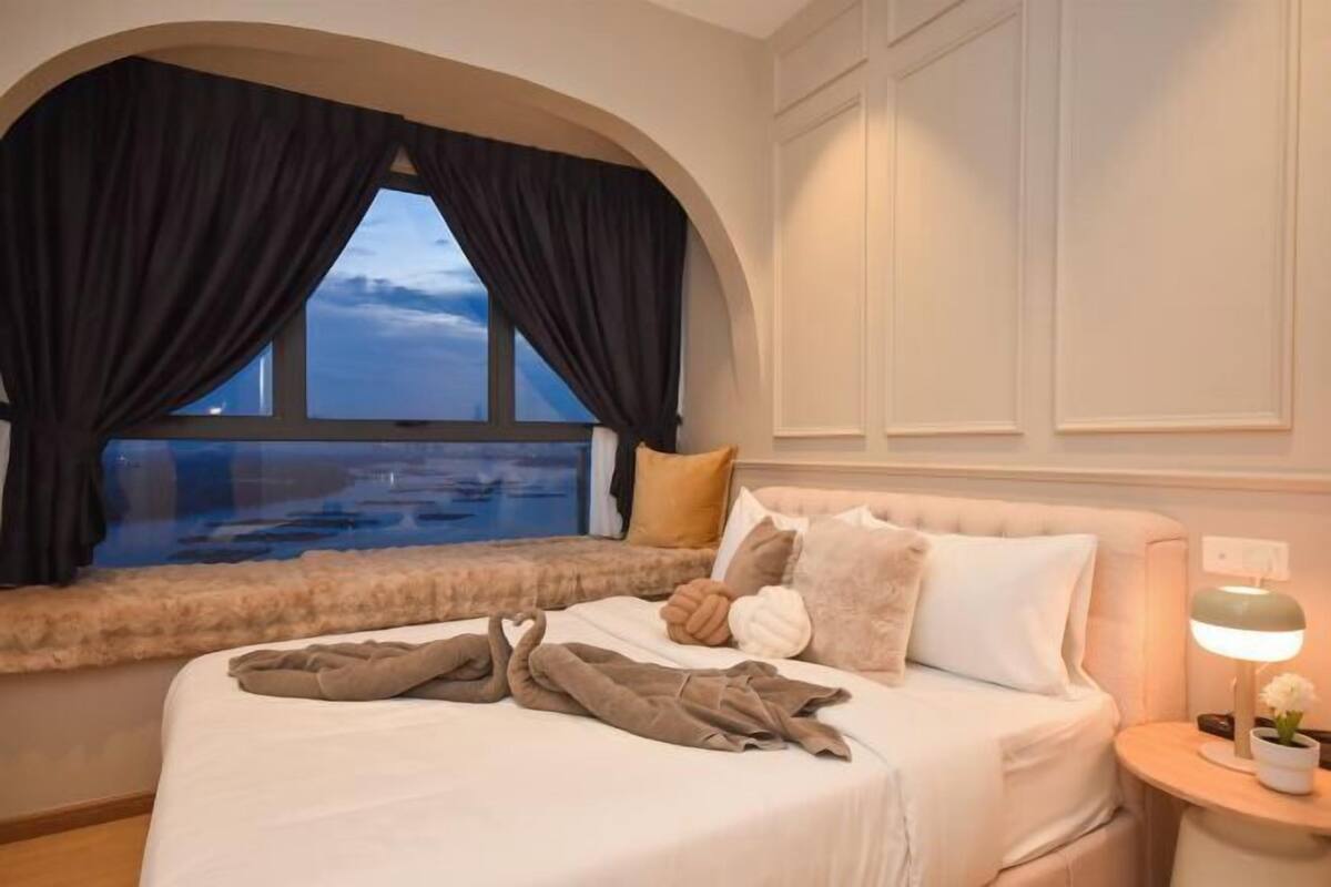 Quarto de hotel com uma janela grande, uma cama com travesseiros e cobertor, uma mesa de cabeceira com abajur e vista para a água.