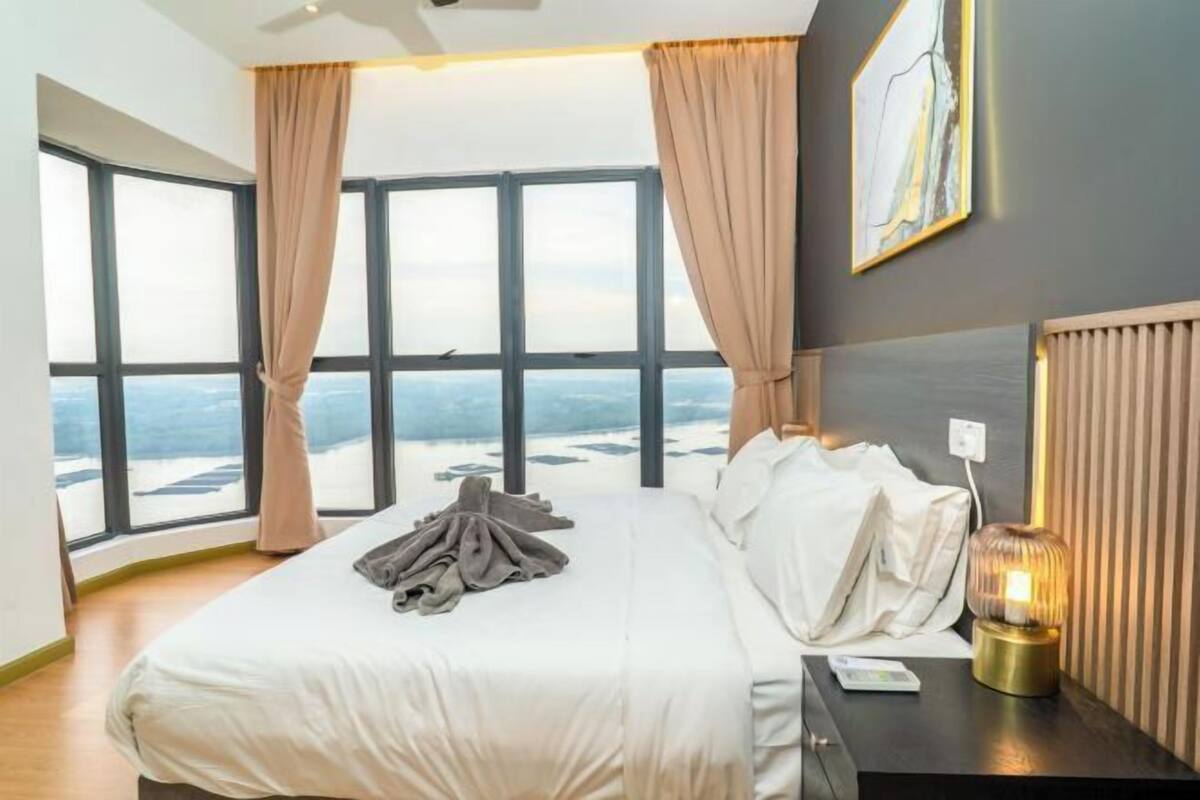 Quarto com uma cama grande, uma mesa de cabeceira, uma luminária e vista do céu através de grandes janelas.