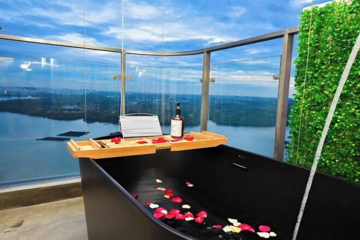 Uma banheira envidraçada com pétalas de rosa e uma garrafa, cercada por uma parede verde e vista para a cidade.