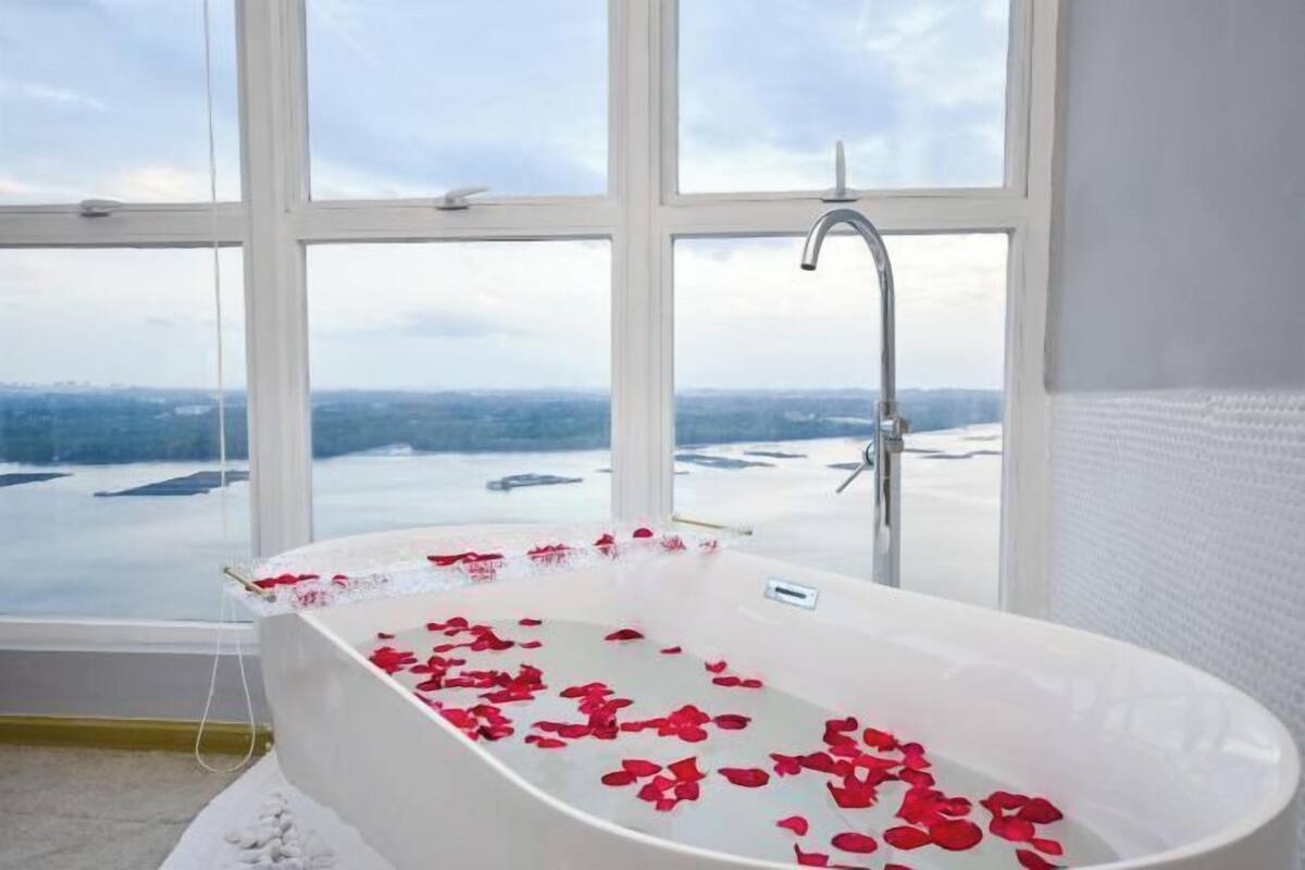 Uma banheira com pétalas de rosa, uma janela com vista para um corpo de água e uma parede com azulejos brancos.