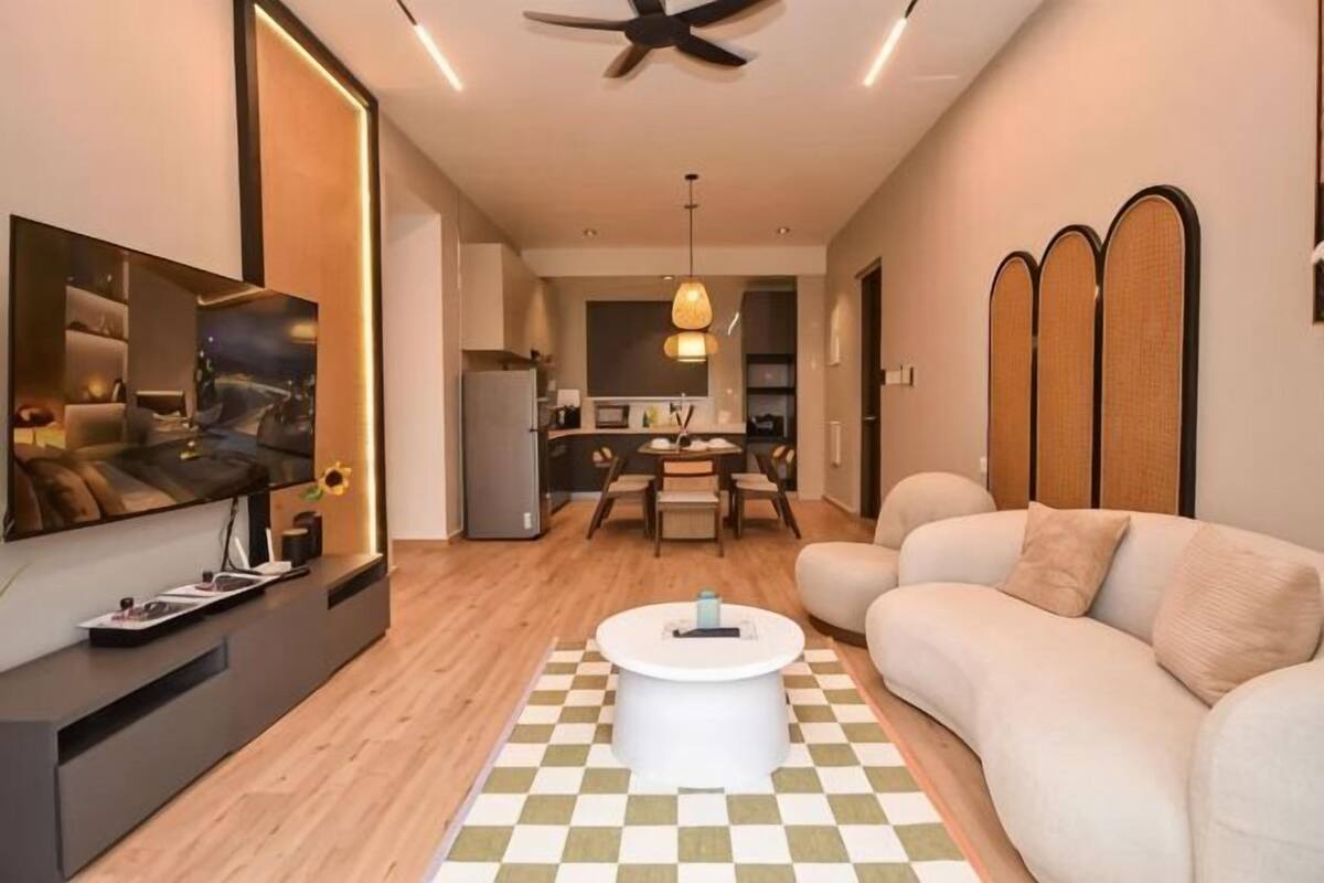 Uma sala de estar moderna com um sofá, uma mesa de centro e uma televisão. Uma cozinha visível ao fundo.